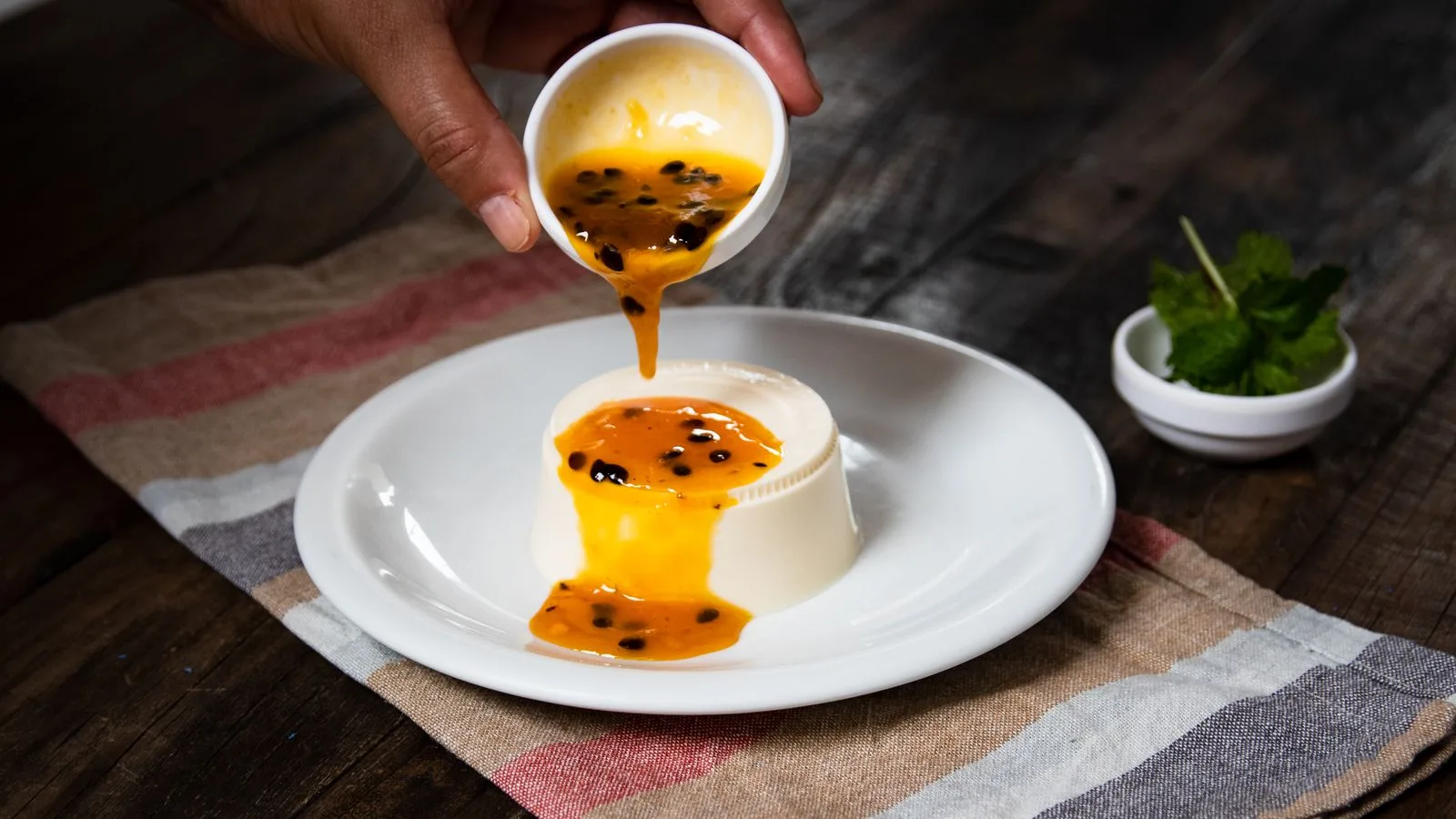 Close-up of a hand pouring passion fruit sauce over panna cotta on a rustic table.