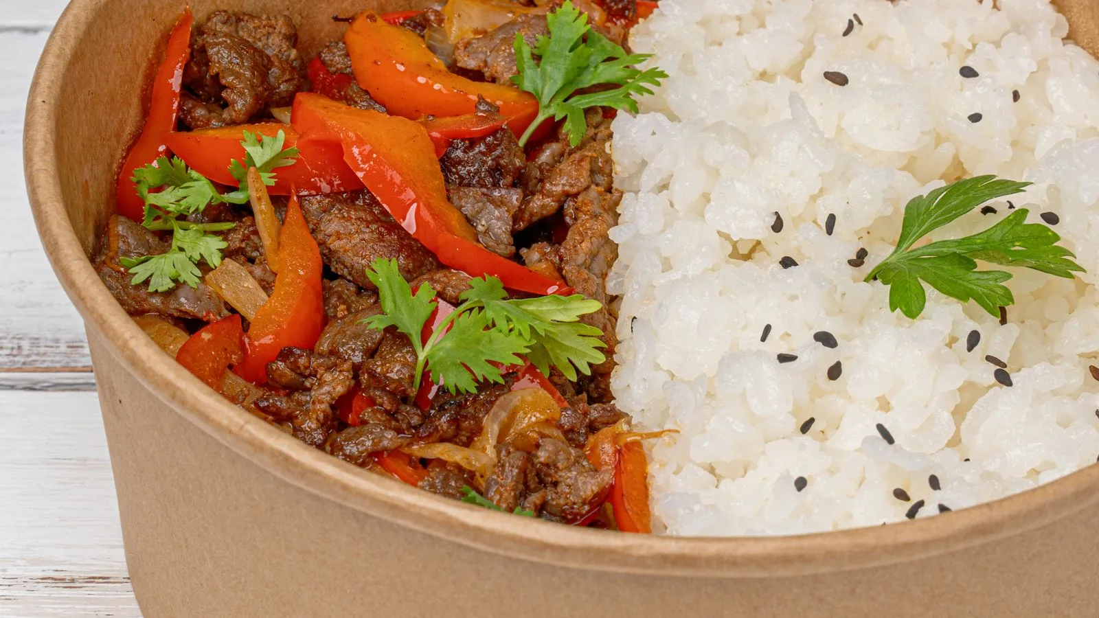 Close-up of a savory beef and vegetables dish with rice in a paper bowl.