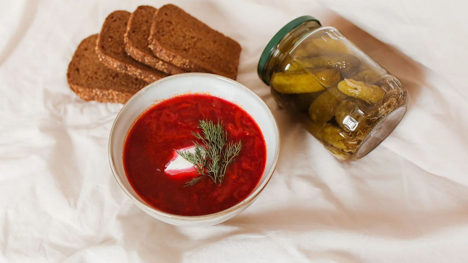 A hearty bowl of borscht with dill, accompanied by sliced brown bread and pickles on a white surface.