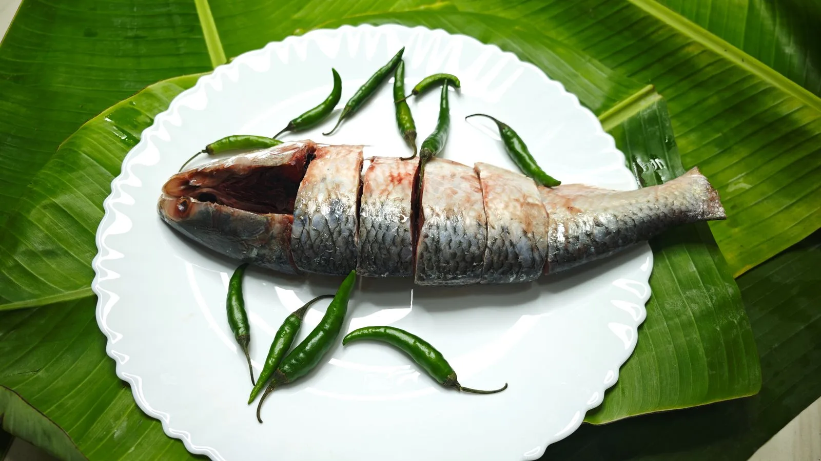 A slice fish garnished with green chilies on a white plate over banana leaves.