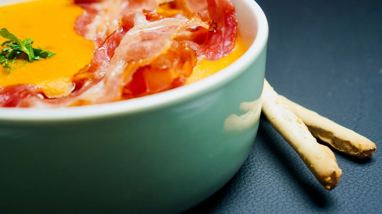 A close-up of a creamy orange soup topped with crispy bacon and herbs in a ceramic bowl (food photography).