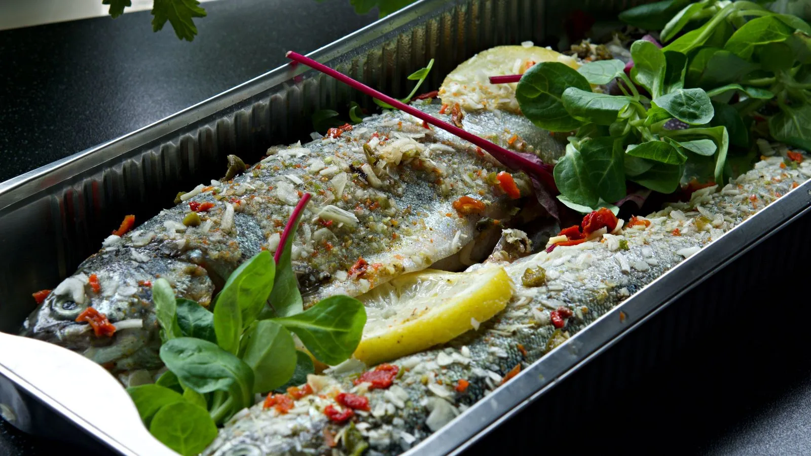 Freshly baked trout in an aluminum tray garnished with lemon, herbs, and spices.
