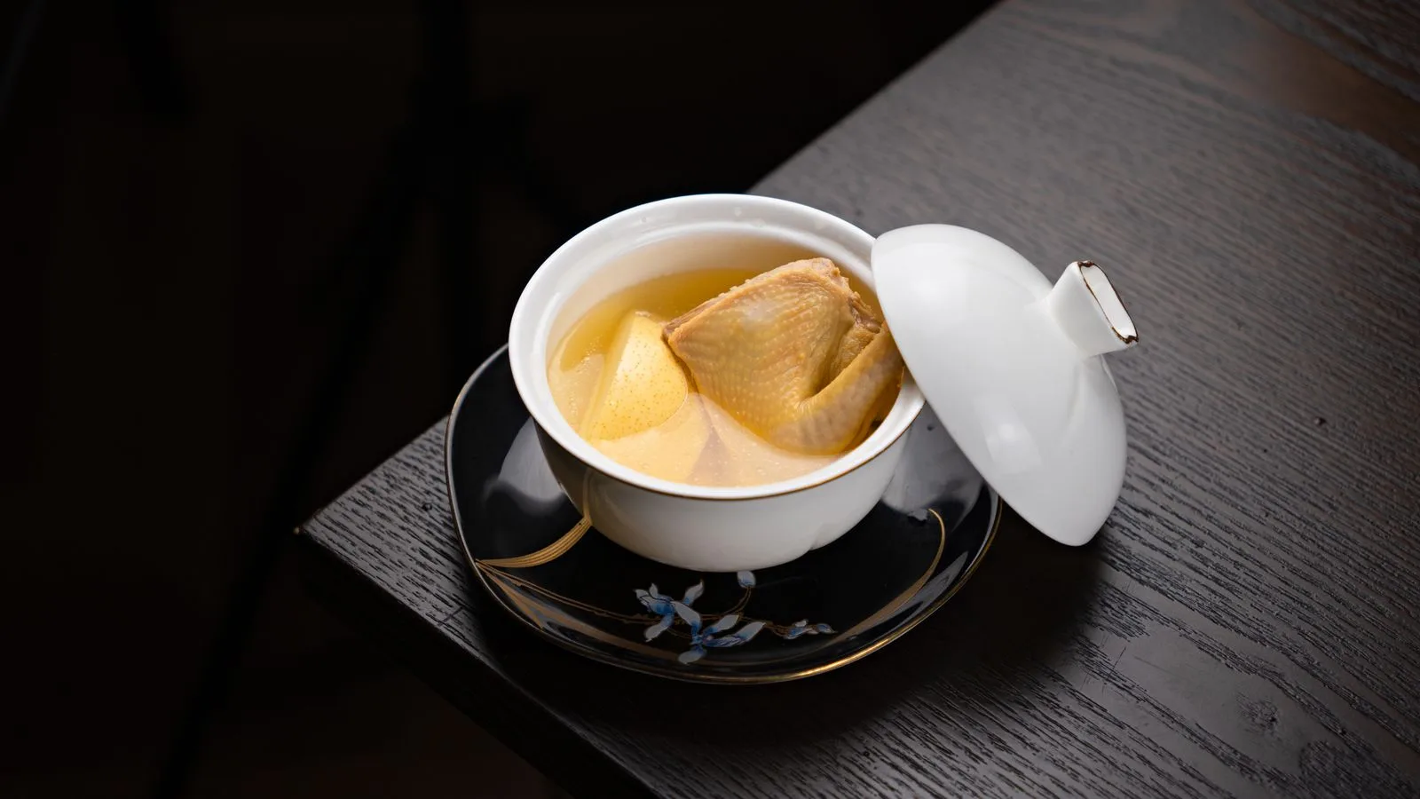 A gourmet bouillon with poultry served in an ornate porcelain bowl on a dark wooden table.