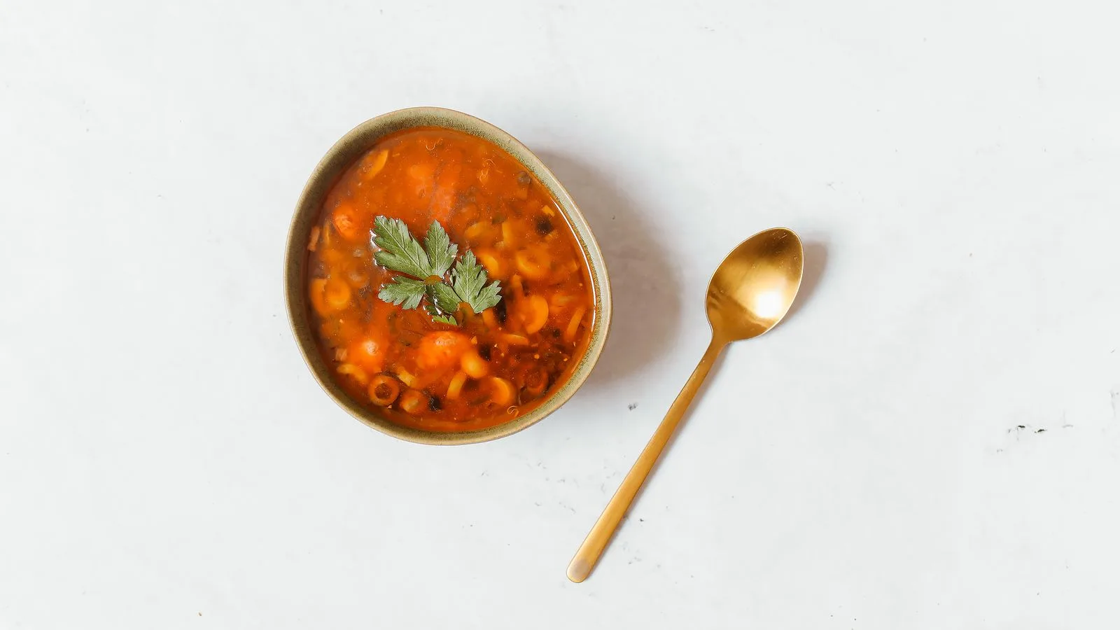 A rustic bowl of Russian solyanka soup with a gold spoon on a white surface.
