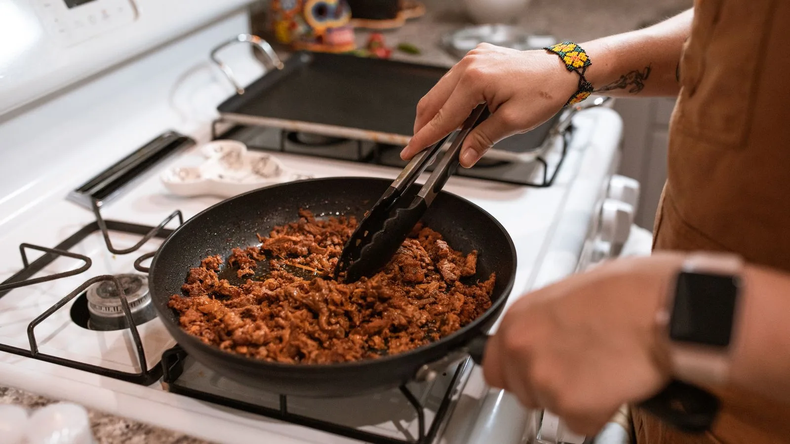Hands cooking taco meat in a pan on a gas stove with tongs.