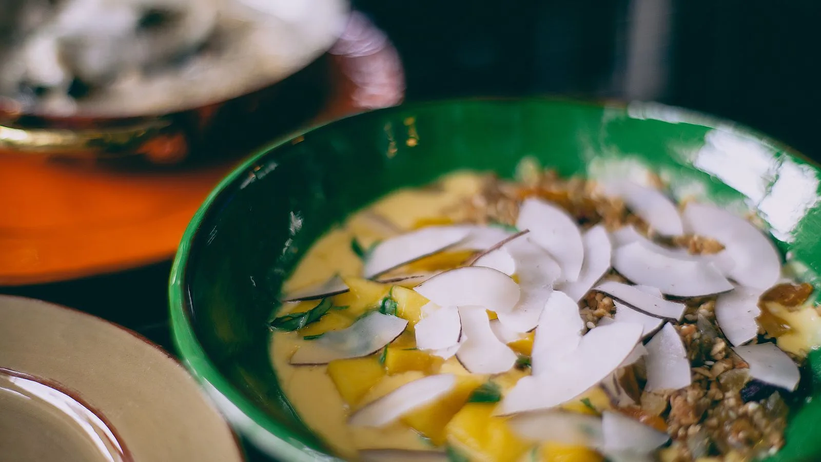Green bowl of tasty homemade soup decorated with slices of coconut placed on table with plates on blurred background in kitchen