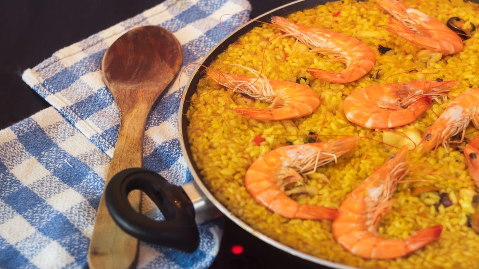 Close-up of a seafood paella with prawns in a pan, a wooden spoon, and blue towel.