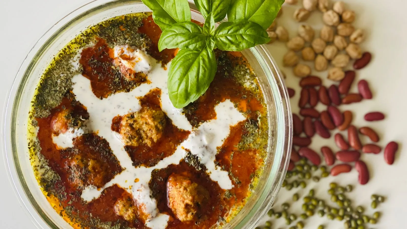 A mouth-watering meatball soup topped with basil leaves, surrounded by colorful beans.