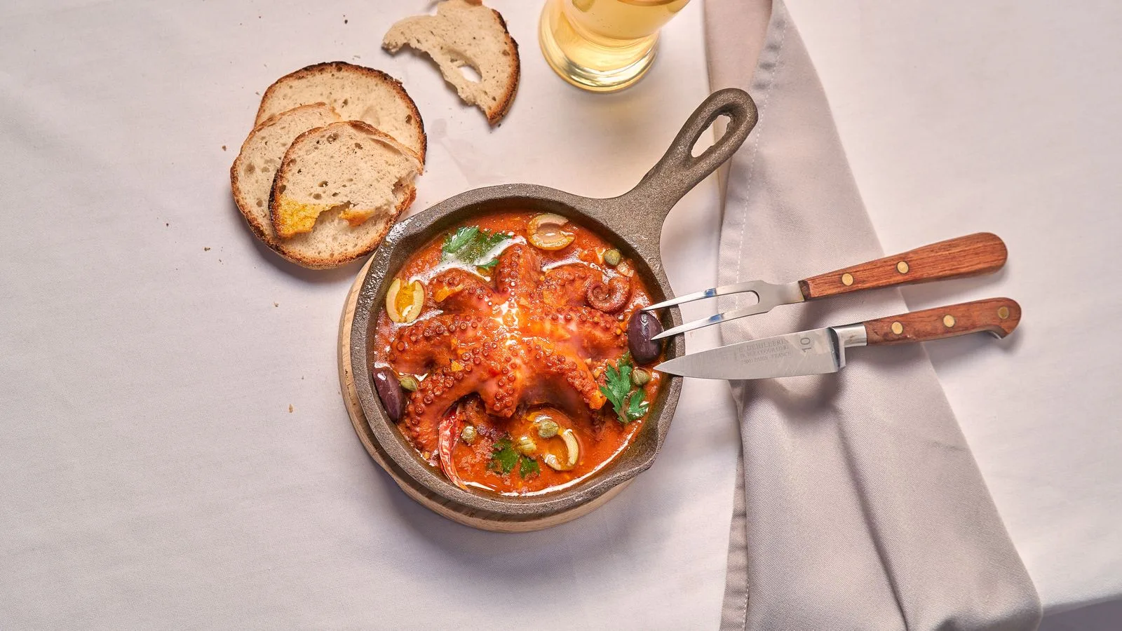 Grilled octopus in a pan with sliced bread, olives, and garnish, ready to be served.