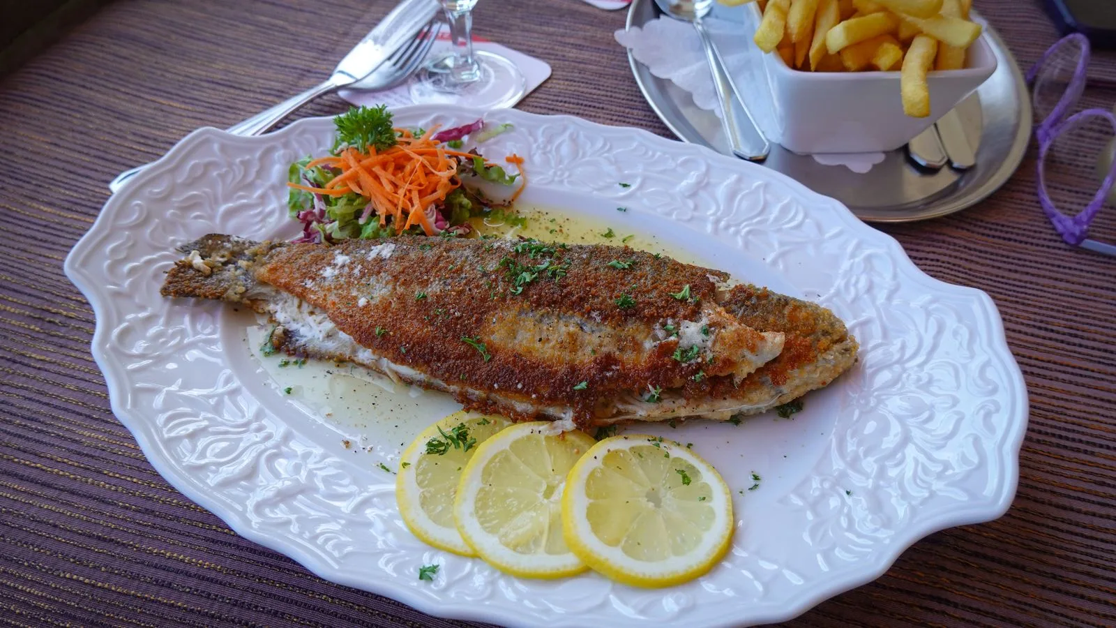 Grilled fish served with lemon slices, fries, and a side salad on a decorative plate.